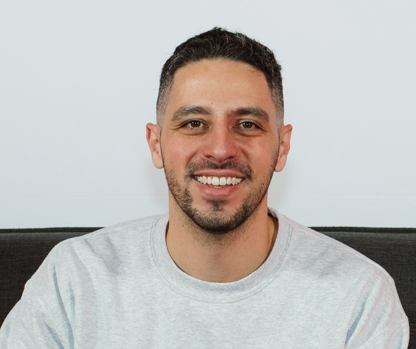 Man with dark hair and beard wearing light gray t-shirt smiling warmly.