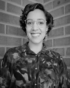 Portrait of person with glasses and colorful blue and purple patterned shirt against brick background.