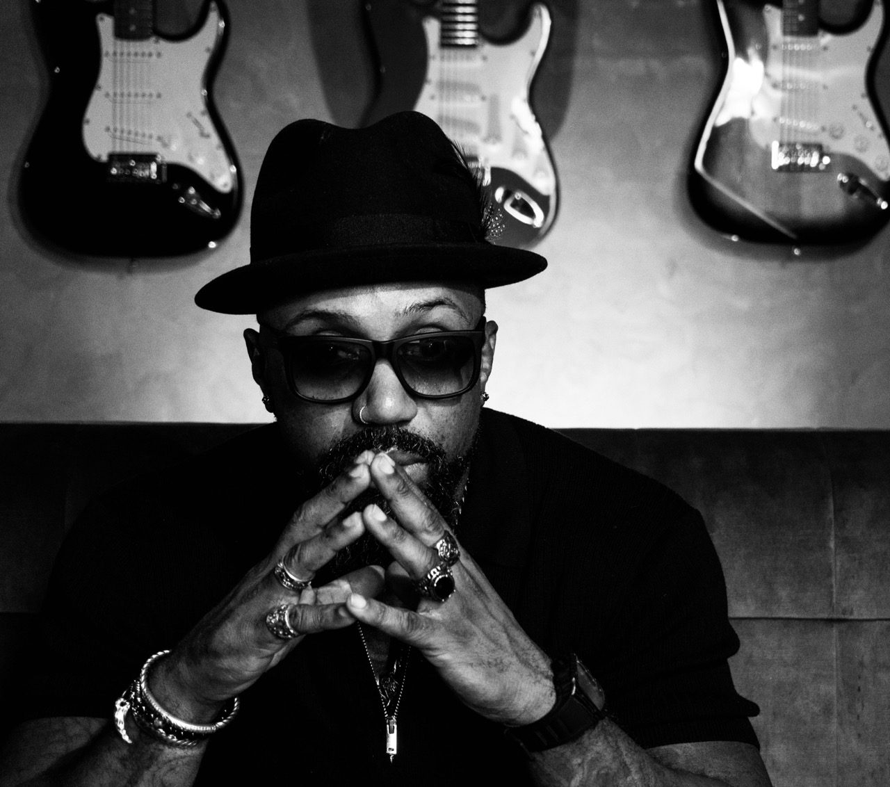 Man with beard and glasses wearing black, black and white portrait with guitars in background.