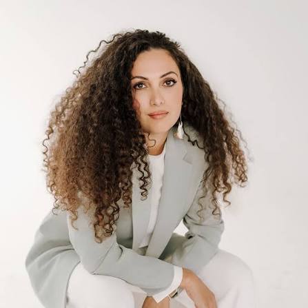 Woman with curly dark hair wearing purple turtleneck with colorful necklace.